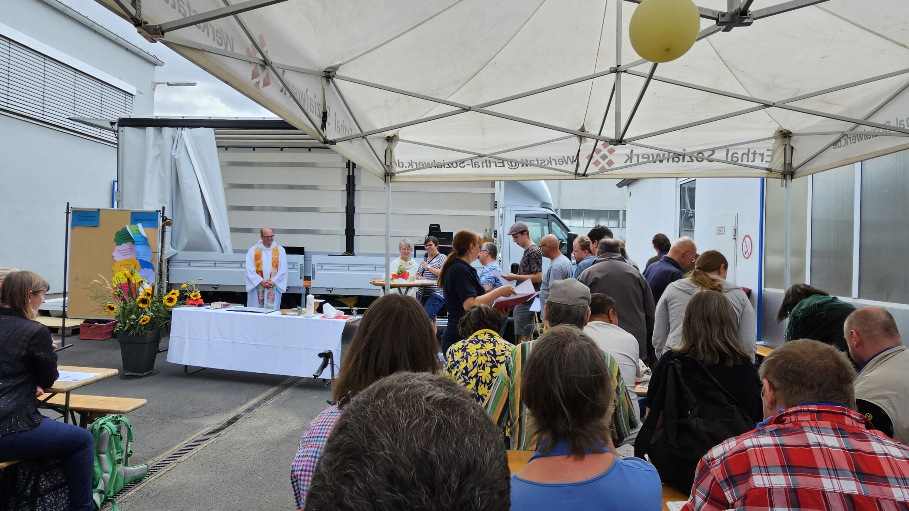 Gruppe von Menschen sitzen auf Bänken während des Freiluftgottestdienstes anlässlich des 25-jährigen Jubiläums des Standorts Heidingsfeld der WfbM des Erthal-Sozialwerks.