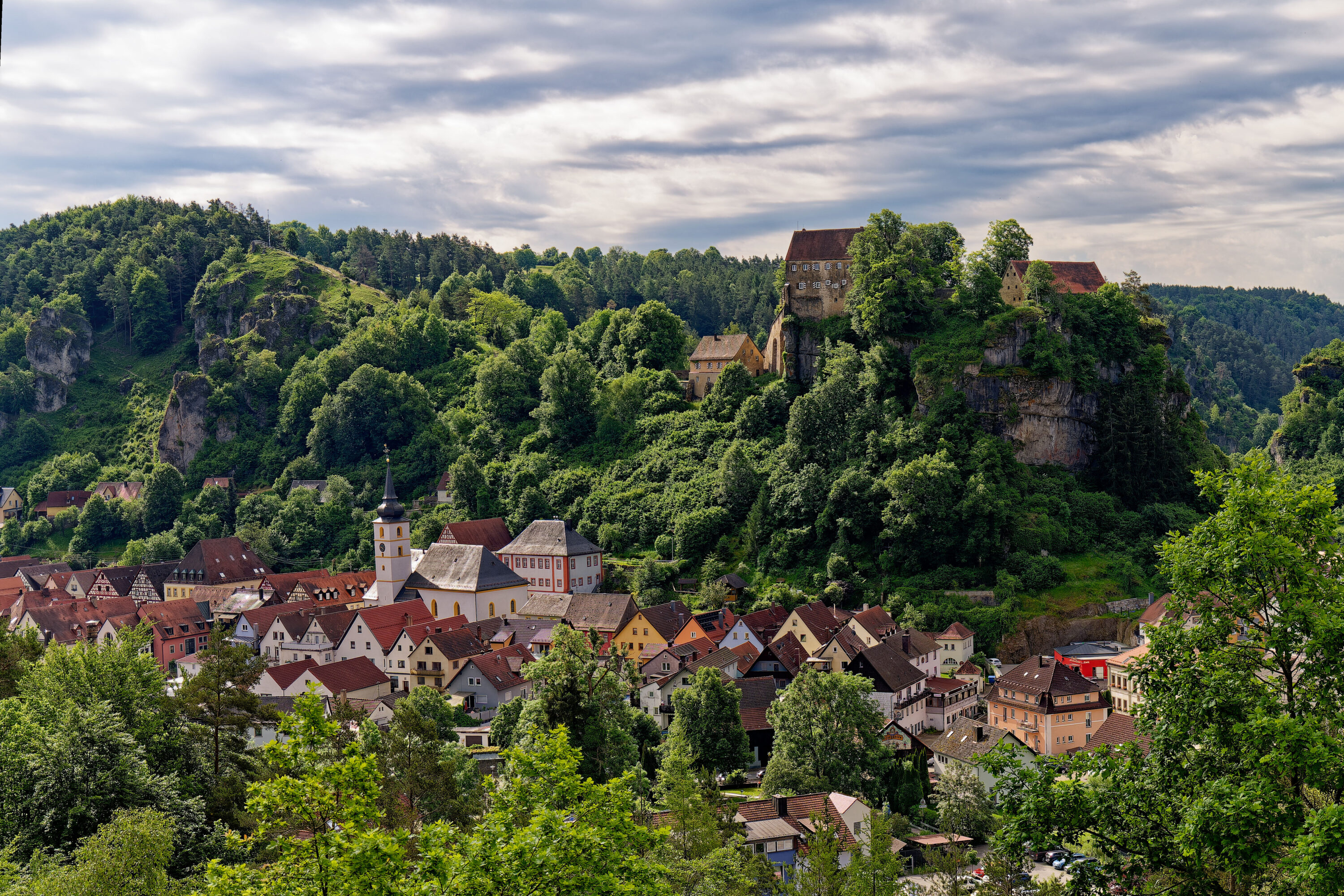 Blick auf eine idyllische fränkische Kleinstadt mit Fachwerkhäusern, Kirche und einer Burg auf einem bewaldeten Felsen.
