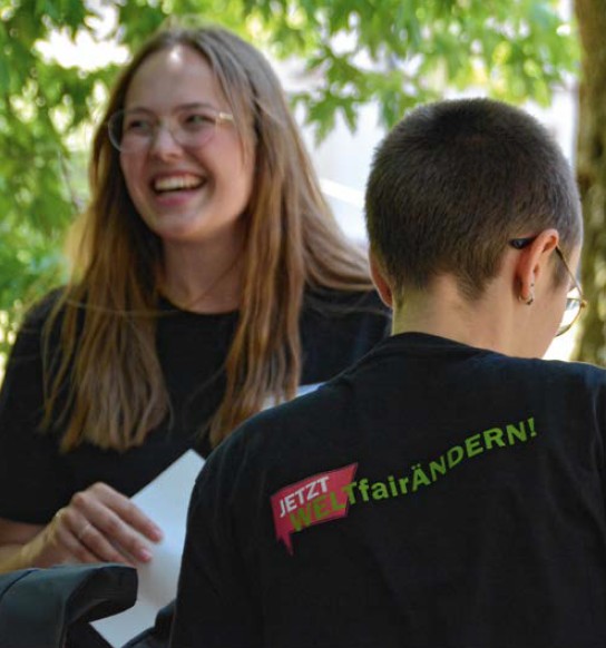 Eine lachende junge Frau steht unter einem Baum. Eine zweite Person steht mit dem Rücken zum Fotografen. Auf dem T-Shirt der zweiten Person steht "Jetzt Welt fairändern".