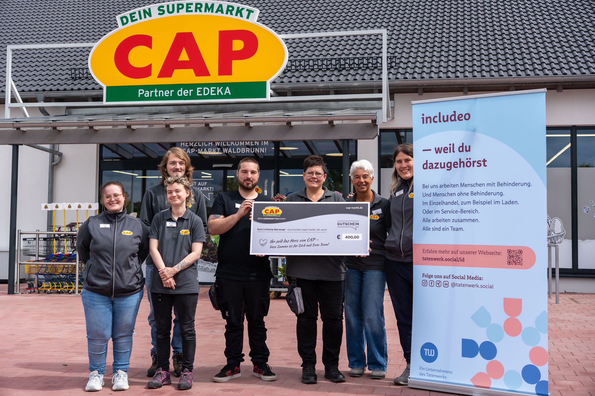 Das Team des CAP-Markts Waldbrunn steht vor dem Eingang des Supermarkts und hält einen Gutschein über 400 Euro für hervorragende Kundenbewertungen in die Kamera. Rechts daneben steht ein Roll-up des inklusiven Unternehmens includeo.