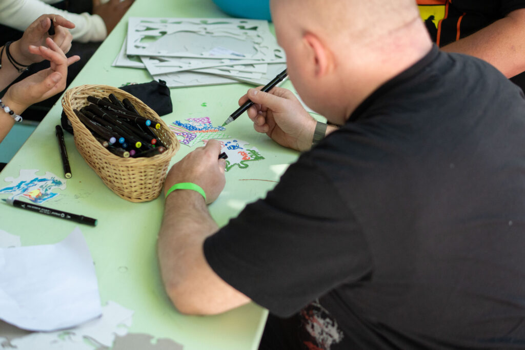Ein Mann malt mit einem Filzstift ein buntes Motiv auf Papier. Auf dem Tisch liegen Schablonen, Stifte und Bastelmaterialien, während weitere Personen gemeinsam kreativ arbeiten.