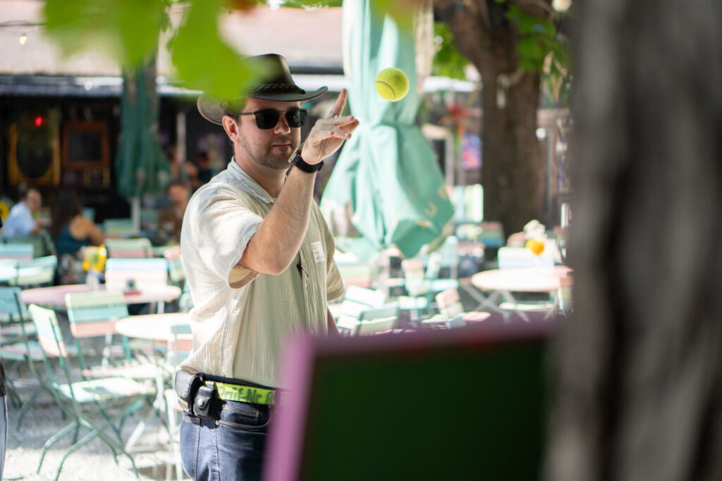 Ein Mann wirft einen Tennisball auf ein Spielziel im Biergarten der Waldschänke Dornheim. Im Hintergrund sitzen Besucher:innen der Dornklusion an Tischen unter schattigen Bäumen.