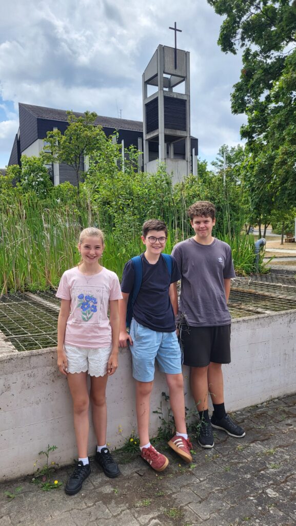 Florentine Götz, Leopold Götz und Philipp Grohmann stehen lächelnd vor dem Teich auf dem Gelände des St. Josefs Stifts. Im Hintergrund sind die Kapelle und der Glockenturm zu sehen. Die Jugendlichen unterstützten beim Vielfalter-Nachmittag die Bewohner:innen bei den verschiedenen Sinnesstationen.