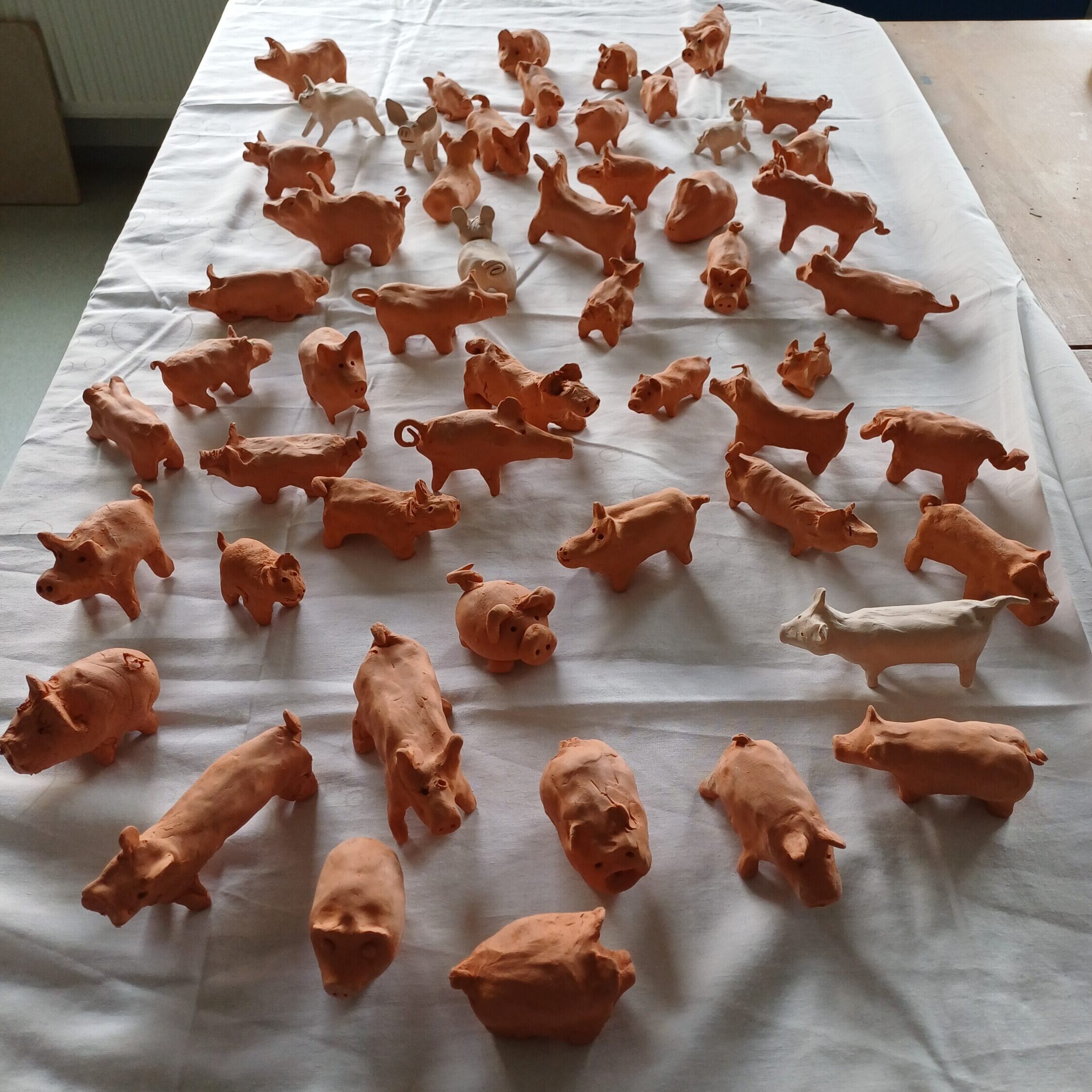 53 unterschiedlich gestaltete Tonschweine stehen auf einem mit einem weißen Tuch bedeckten Tisch. Die Figuren wurden von Schülerinnen und Schülern der Dr. Maria-Probst-Schule für das Kunstprojekt „Schwein für Schwein“ modelliert.