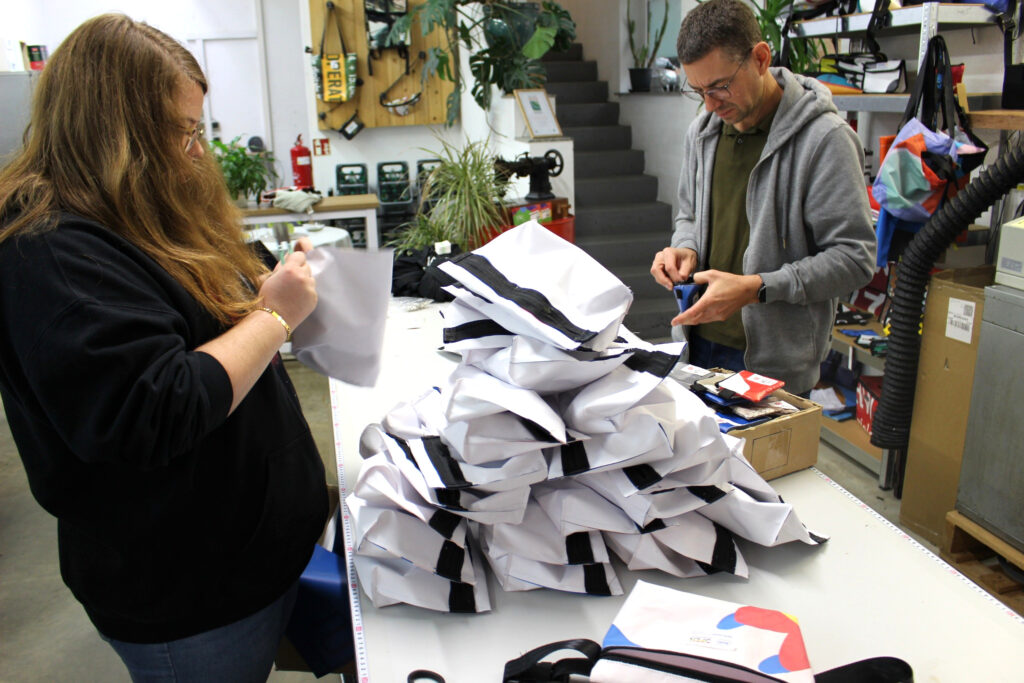 Zwei Personen bearbeiten Taschen und Zubehör an einem Arbeitstisch bei Reciclage. Zwischen ihnen liegen weiße Taschenteile mit schwarzen Reißverschlüssen.