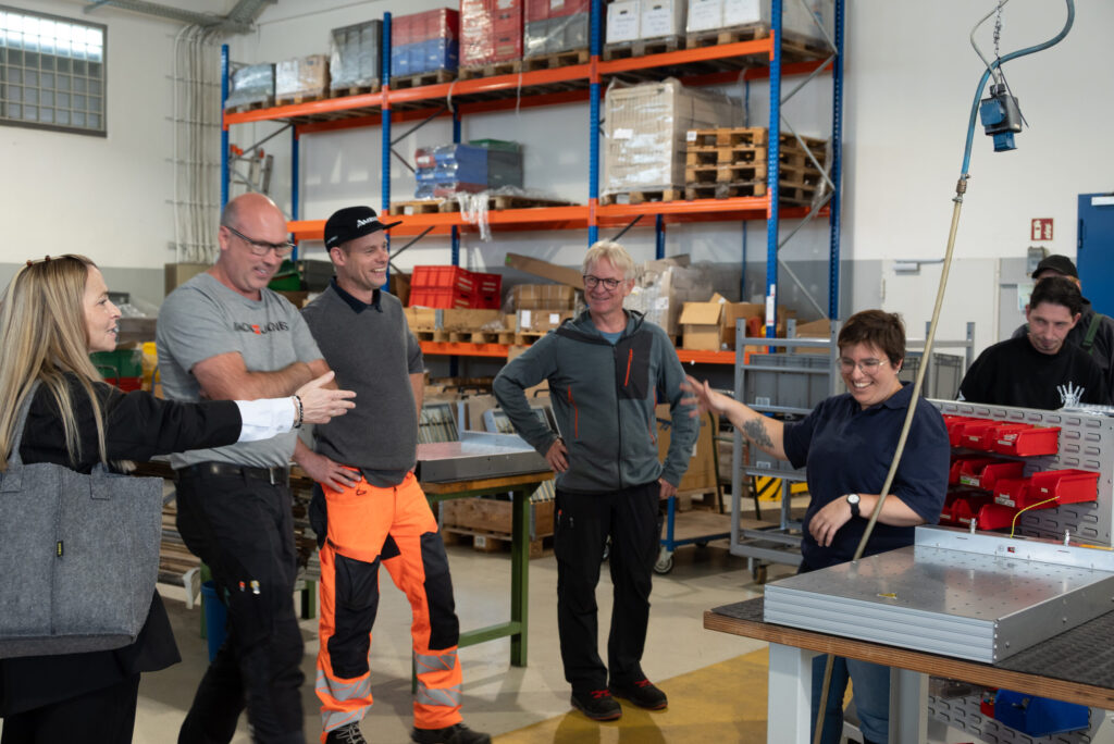 Eine Mitarbeiterin zeigt Gästen einen Arbeitsplatz in der Elektrofertigung. Mehrere Personen lachen gemeinsam.