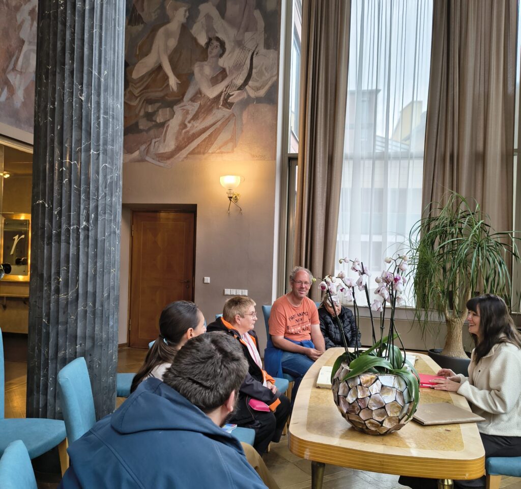 Sieben Personen stehen in der großzügigen Halle des Rathauses und blicken in die Kamera. Über ihnen befinden sich eine große Uhr mit goldenen Zeigern und weitere Zifferblätter. Säulen und offene Galerien prägen den Raum.
