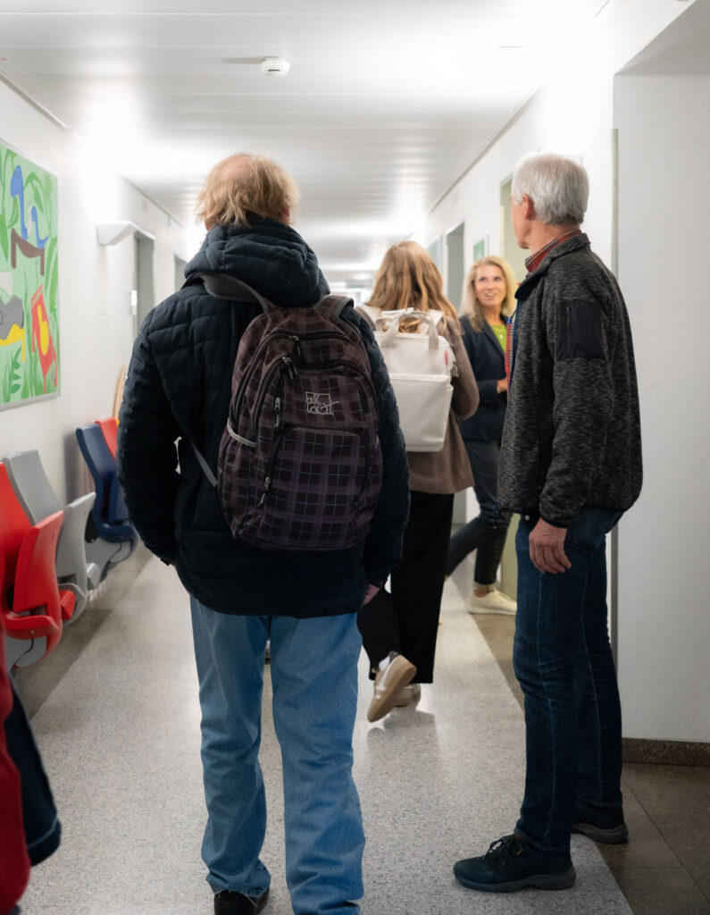 Vier Teilnehmende stehen während eines Rundgangs im Flur der Kinderklinik. An der Wand hängen bunte Bilder, darunter stehen Wartezimmerstühle.
