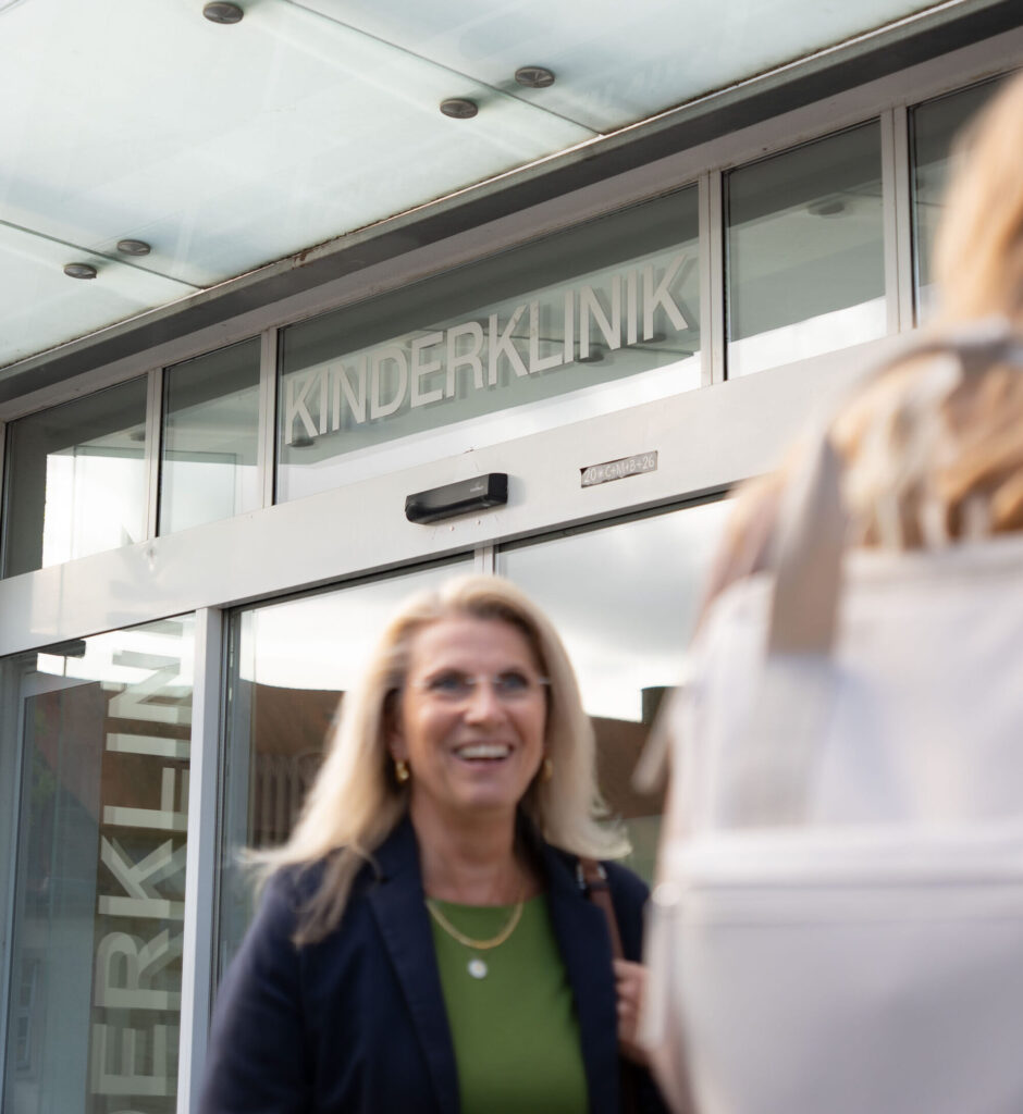 Eine Frau lächelt vor dem Eingang der Kinderklinik Würzburg. Über der Glastür steht „Kinderklinik“.