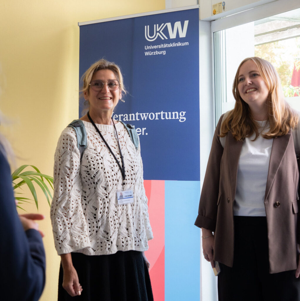 Drei Frauen im Gespräch vor einem Aufsteller des Universitätsklinikums Würzburg. Zwei hören zu, während die dritte gestikuliert.