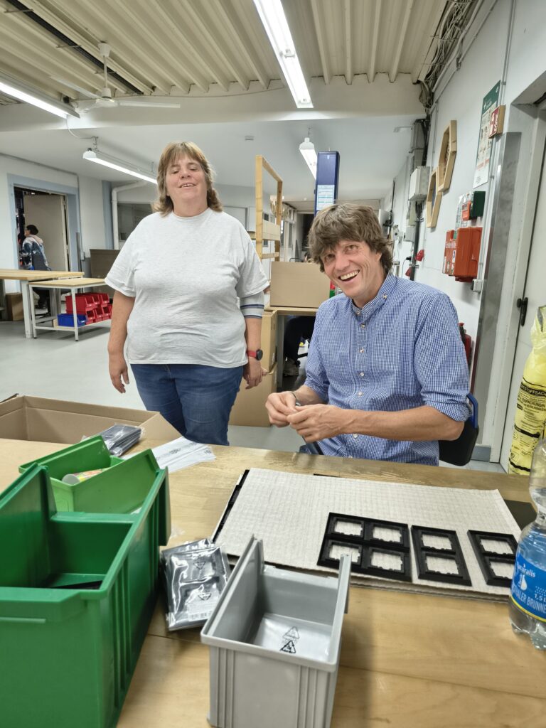 Ein Besucher sitzt lächelnd am Arbeitstisch und bearbeitet ein kleines Teil. Eine Werkstattmitarbeiterin steht neben ihm.