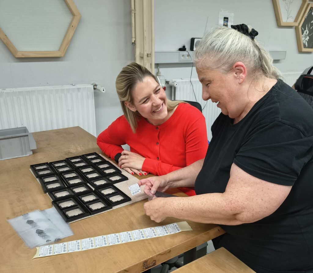 Zwei Frauen sitzen am Arbeitstisch und lächeln einander an. Eine hält ein Etikett und einen transparenten Beutel in den Händen.